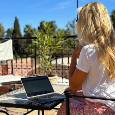 A person with long blonde hair, sits outdoors with a laptop open on a table